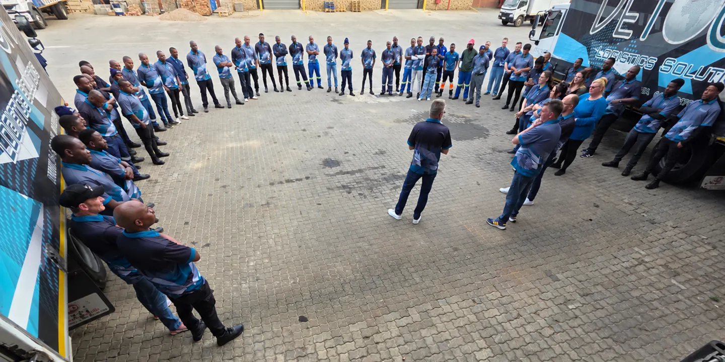 DLS staff in branded shirts stand in a wide circle on the paved yard between two DETOUR-liveried trucks, with one person in the centre facing them.